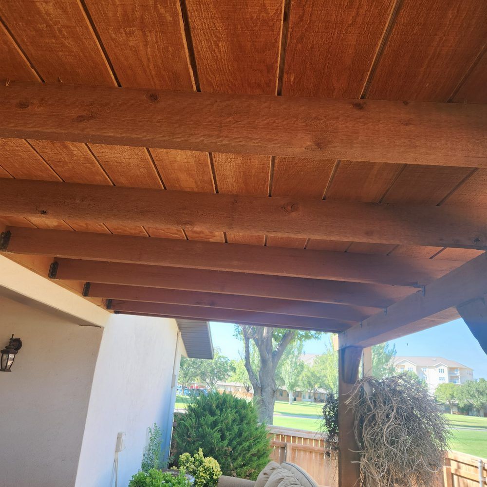 Wooden patio ceiling with beams and planks, view overlooking a green yard and buildings.