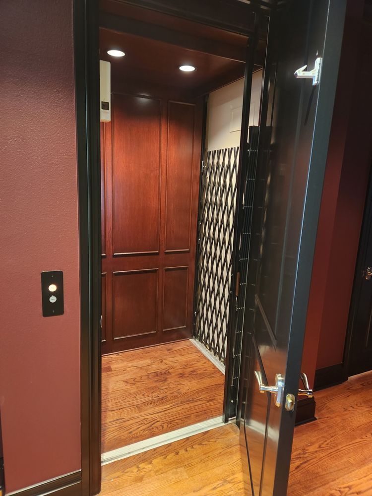 Open residential elevator with cherry-wood interior, patterned wall, and black door with silver handle; maroon wall.