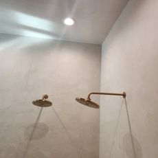 Two gold showerheads in a corner shower with light-colored walls and a recessed ceiling light.