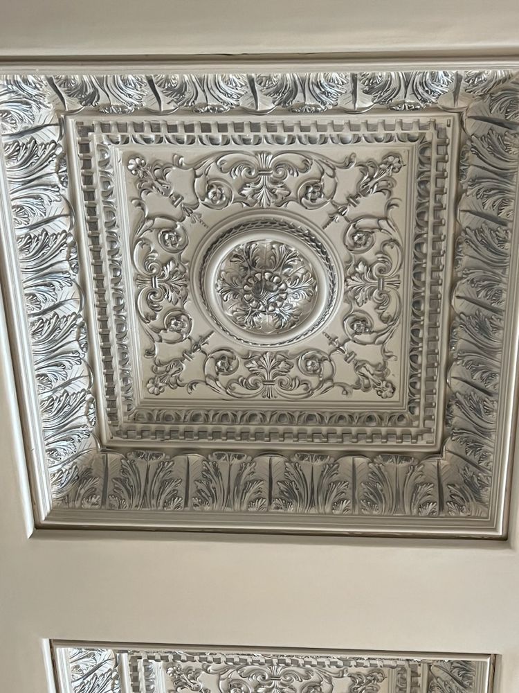 Ornate, white-painted ceiling panel with detailed floral and scroll relief design, centered with a circular medallion.