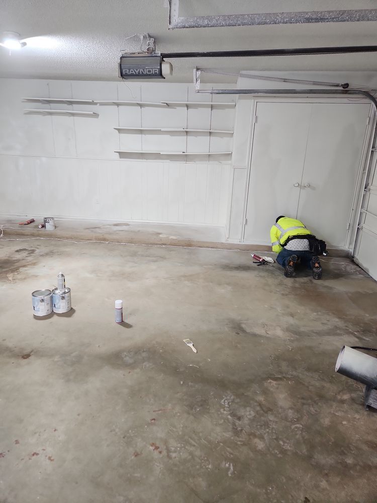 A person kneels in a garage, near paint cans, working on the floor. Shelves are mounted on a wall.