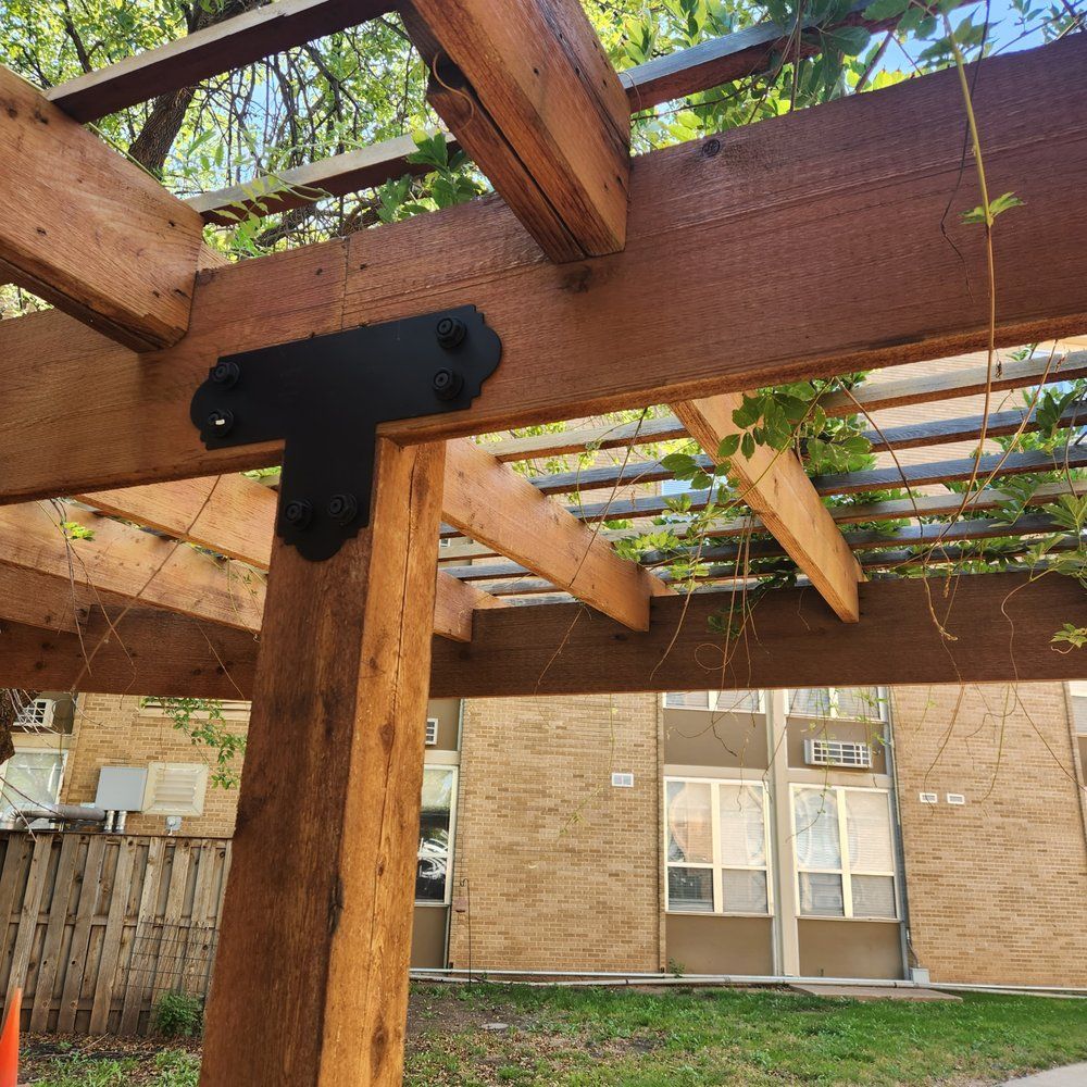 Wooden pergola with black metal brackets, surrounded by greenery, in front of a brick building.