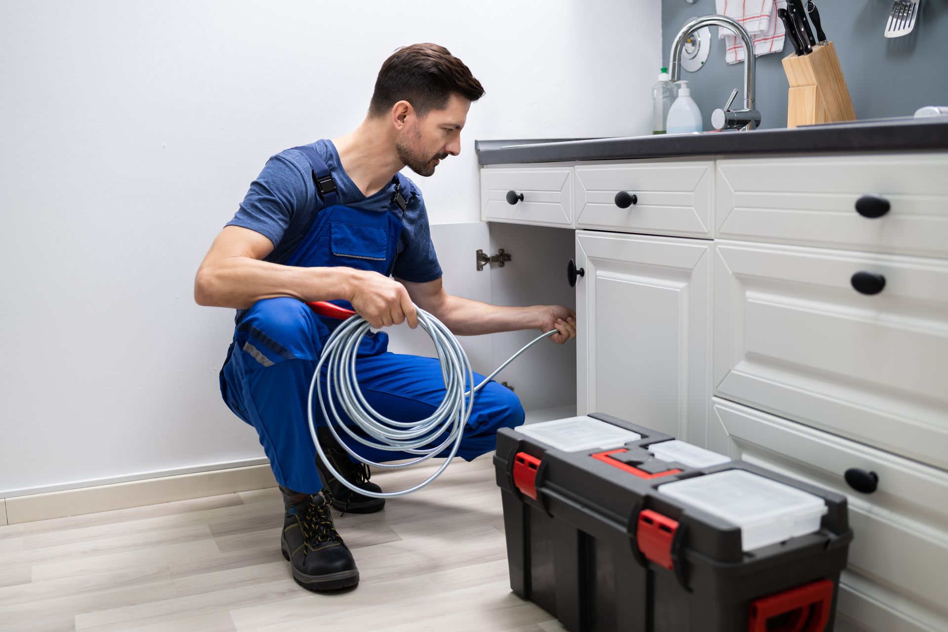 A plumber in blue overalls is cleaning a clogged sink pipe with a hose, a toolbox nearby.