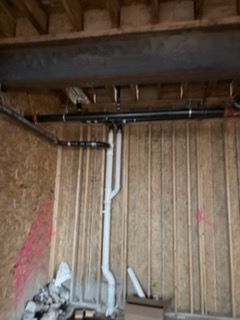 Exposed basement wall with white PVC pipes and rough wooden framing, with debris on the floor.