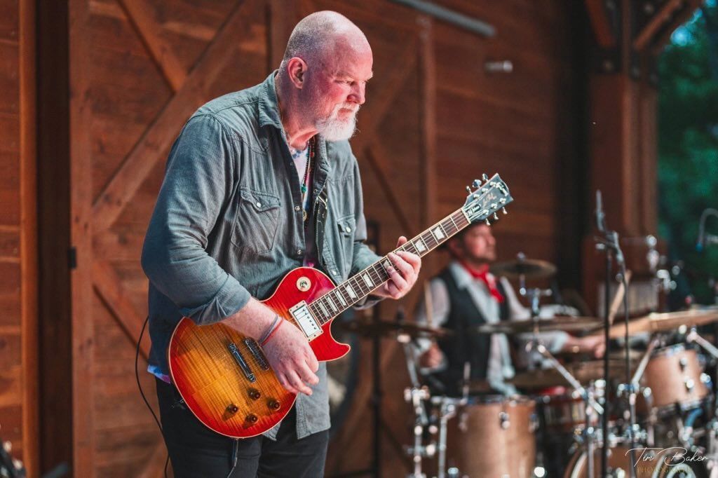 Man with white beard plays red guitar on stage, band in background.