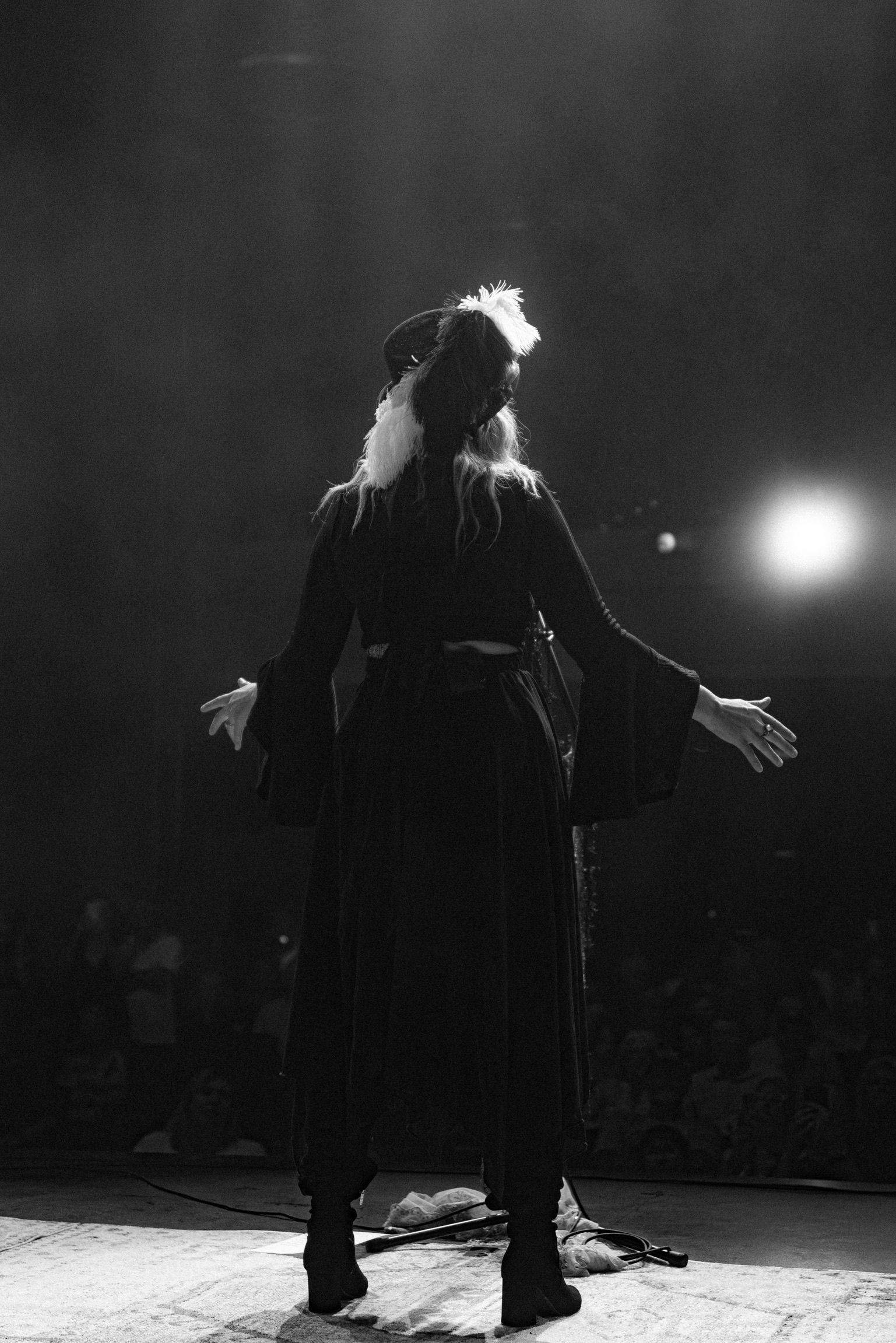 Woman with arms outstretched on a stage, back to camera, wearing dark dress. Stage lit with spotlight.