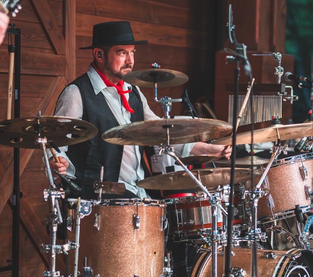Drummer in black hat, vest, and red scarf playing drums in front of a wooden backdrop.