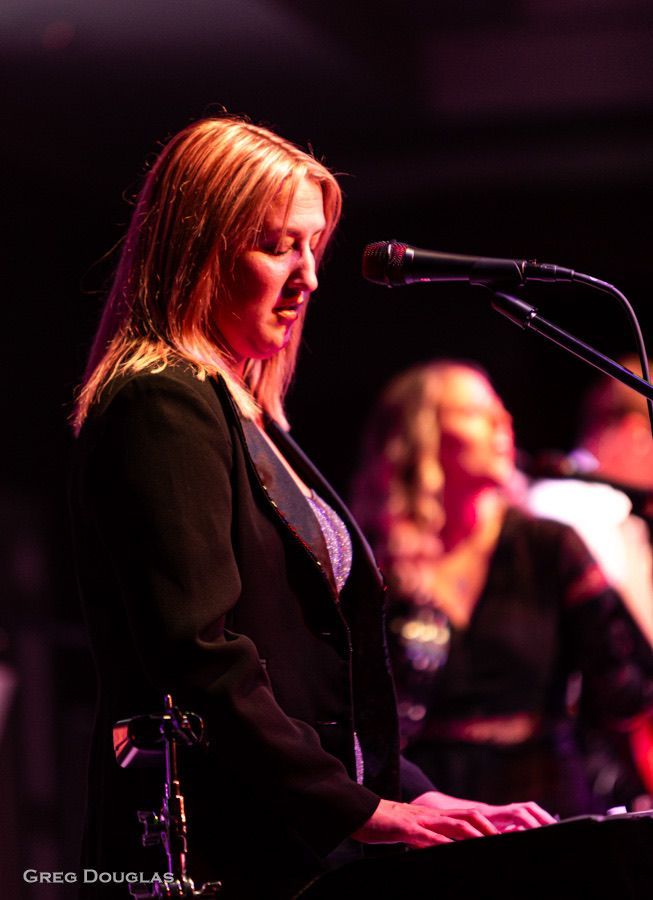 Woman playing keyboard on stage, wearing black jacket, singing into a microphone. Another musician in the background.