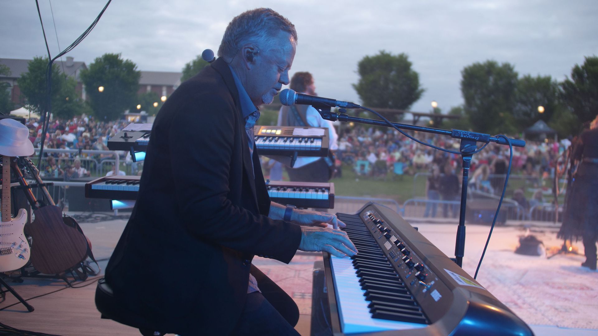 A man in a suit plays a keyboard on an outdoor stage, performing for a crowd.