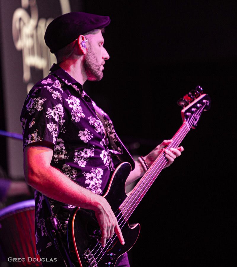 Man playing a black bass guitar on stage, wearing a patterned shirt and cap.