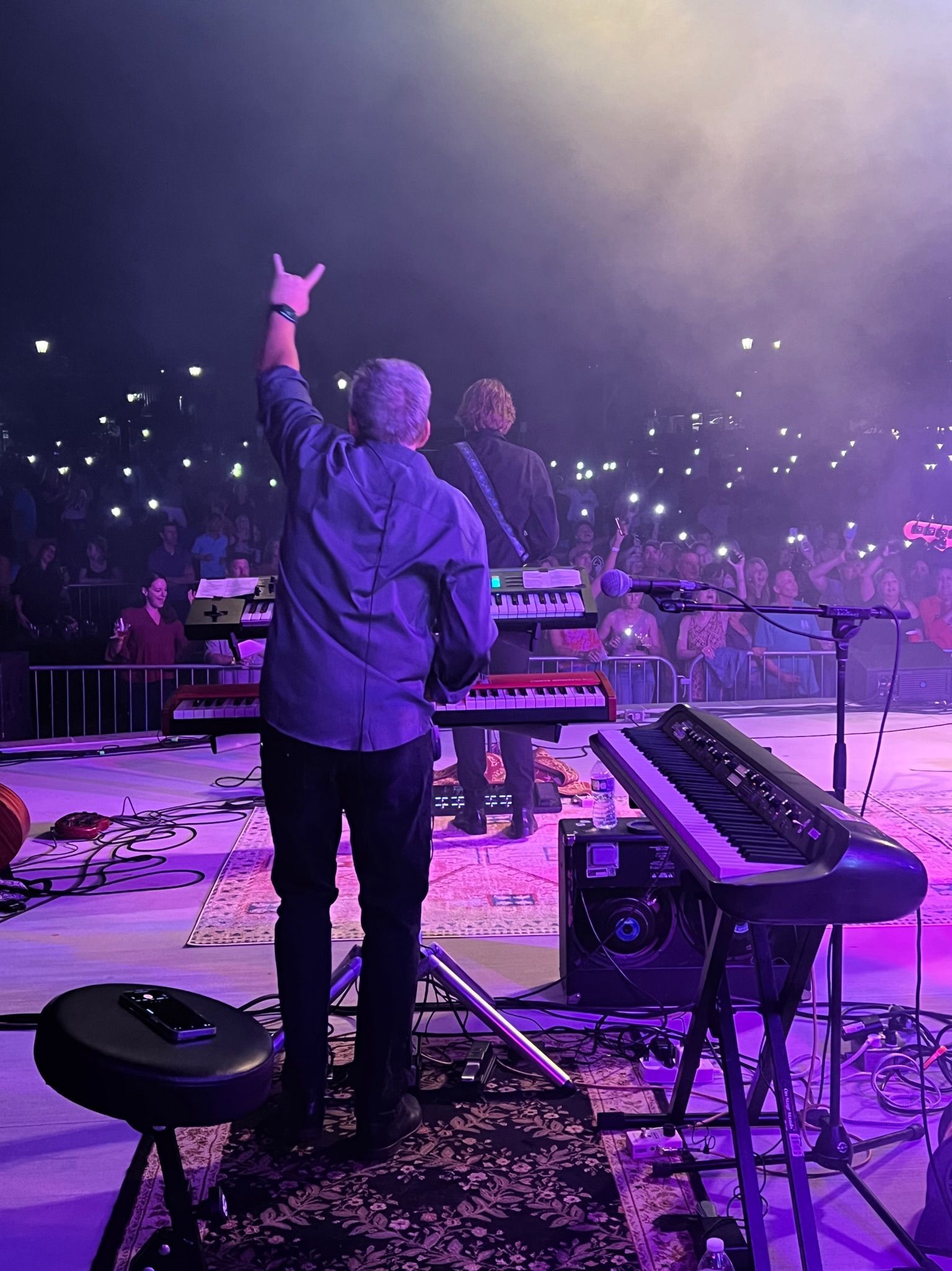 Keyboardist on stage, raising hand in front of a crowd. Purple stage lighting, many lights in background.