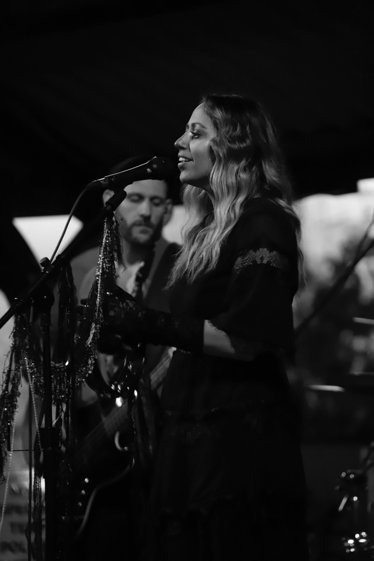 Woman singing at a concert, wearing a black dress, with a band member behind her.