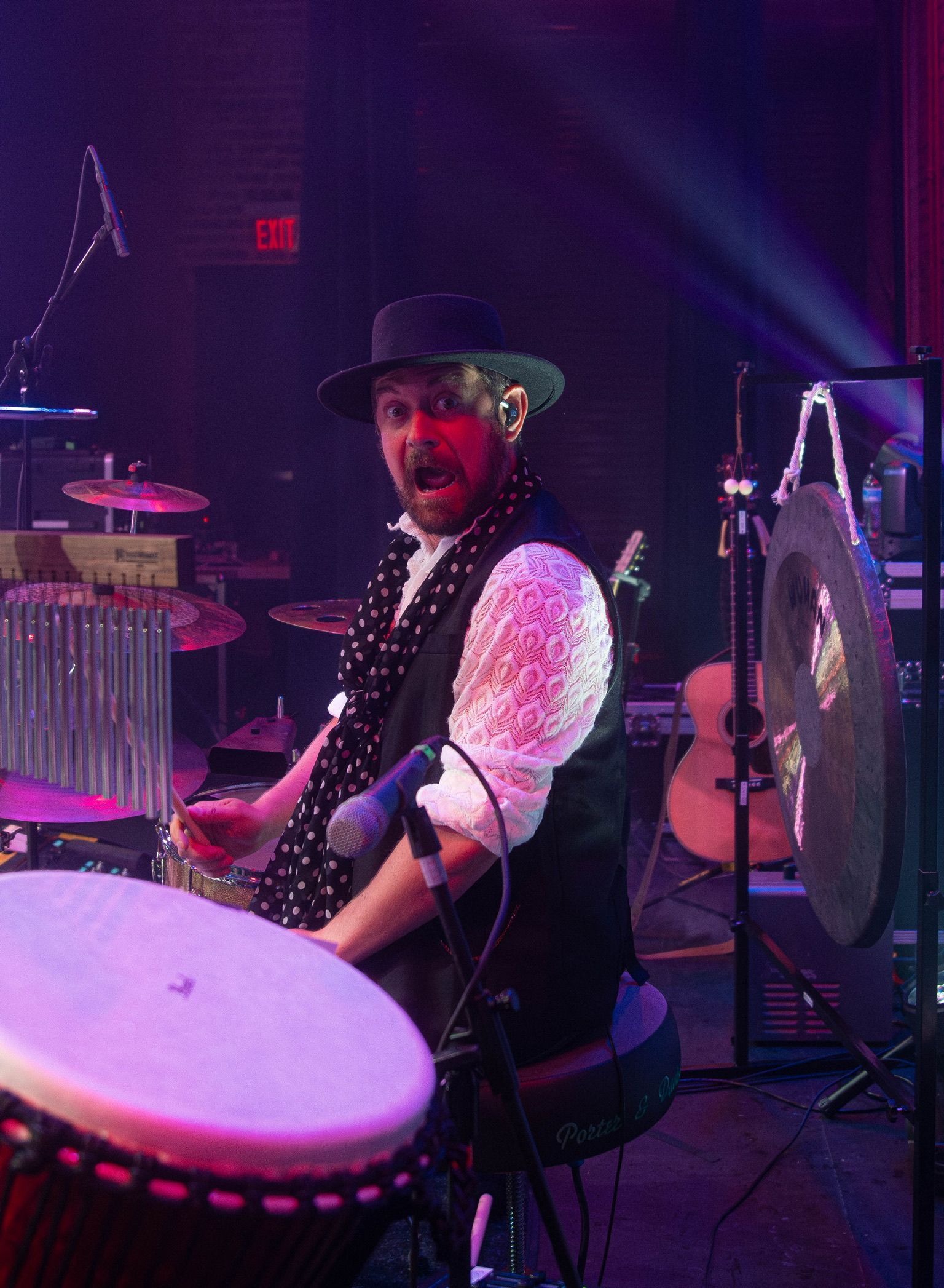 Man playing percussion instruments on stage; wearing black hat, vest, white shirt, and polka-dot scarf.