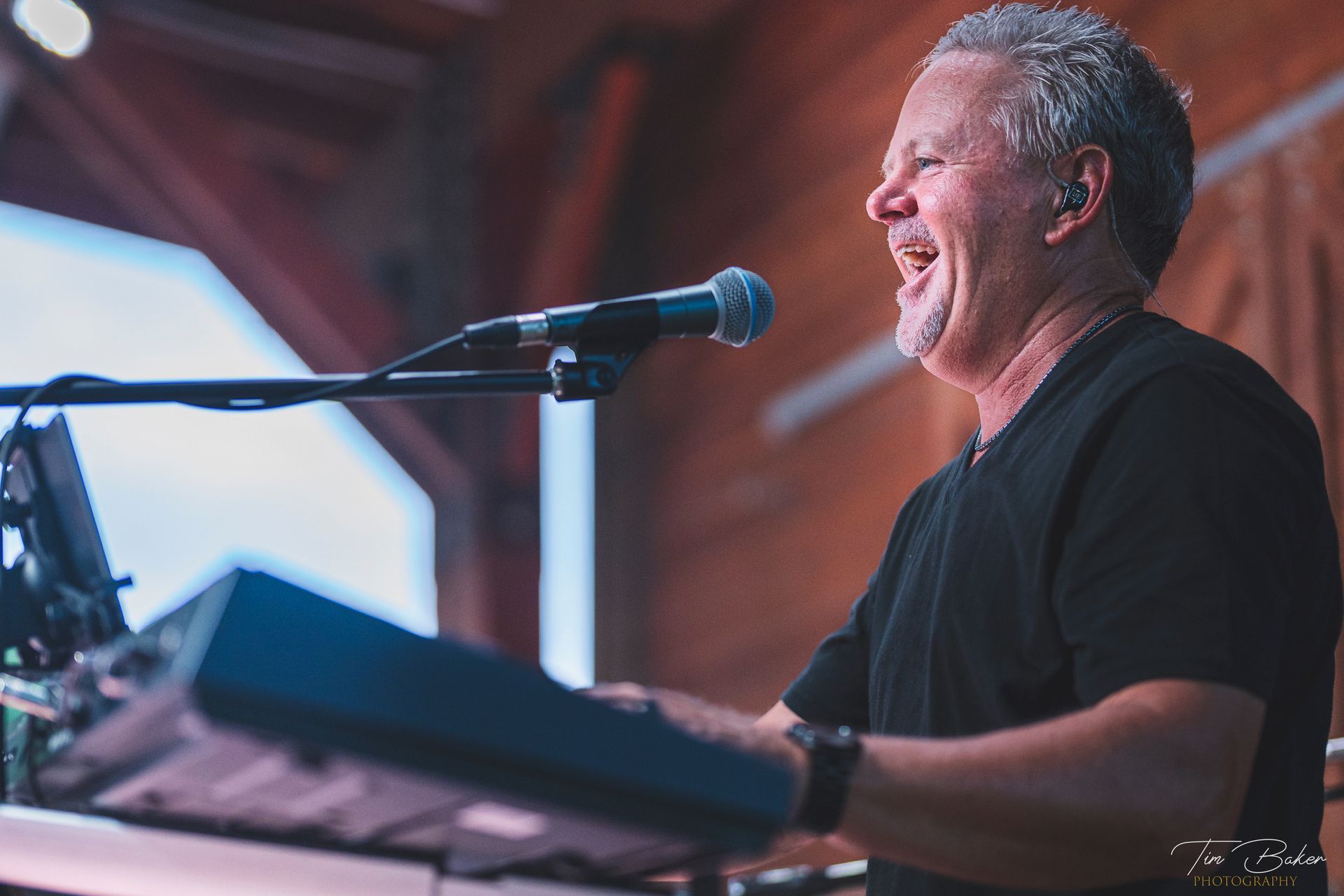 Man singing and playing keyboard on stage, smiling, wearing black shirt.