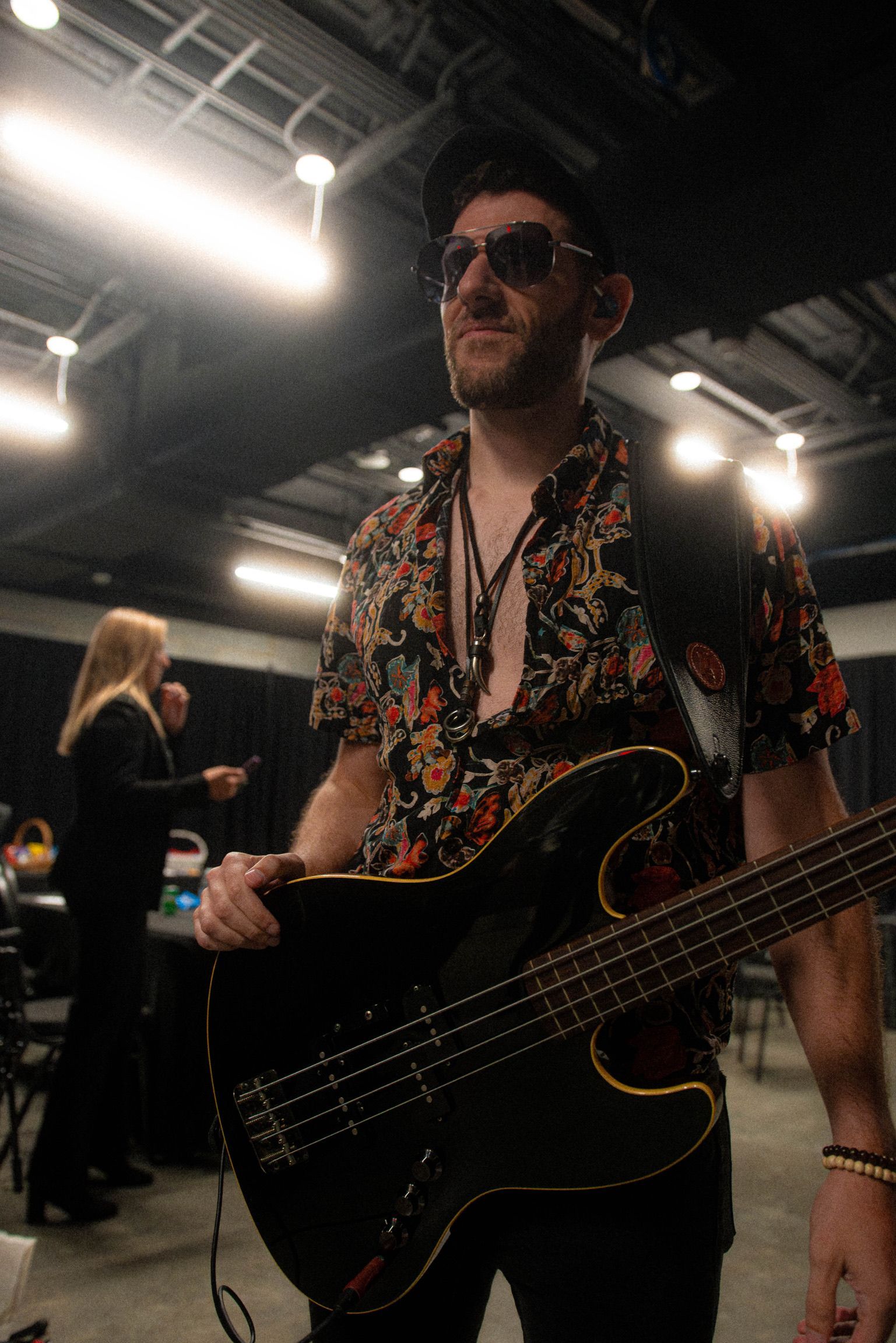 Man holding a black bass guitar, wearing sunglasses, floral shirt, backstage.
