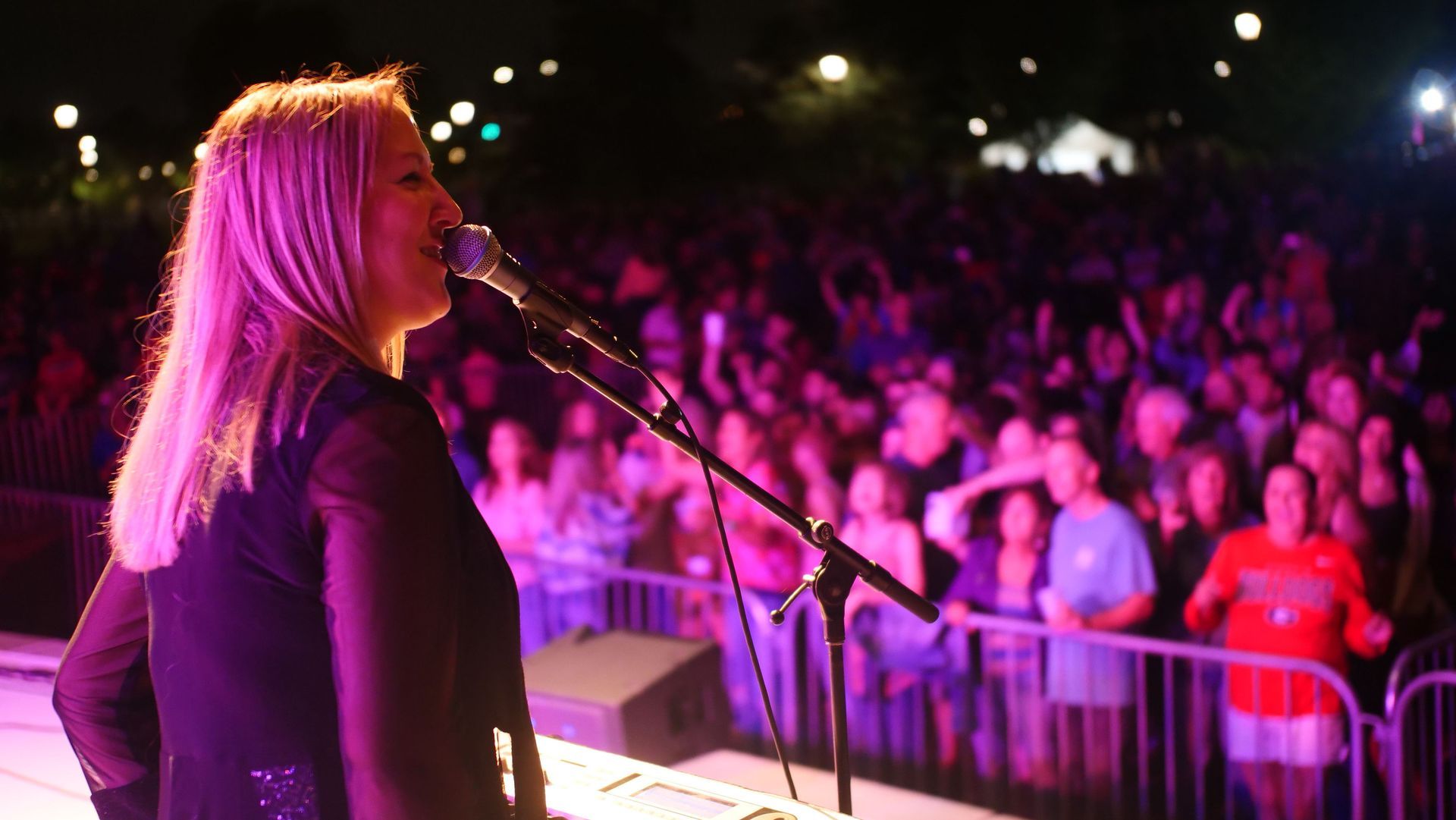 Blonde woman singing into microphone on stage at night, facing a large crowd behind a barrier.
