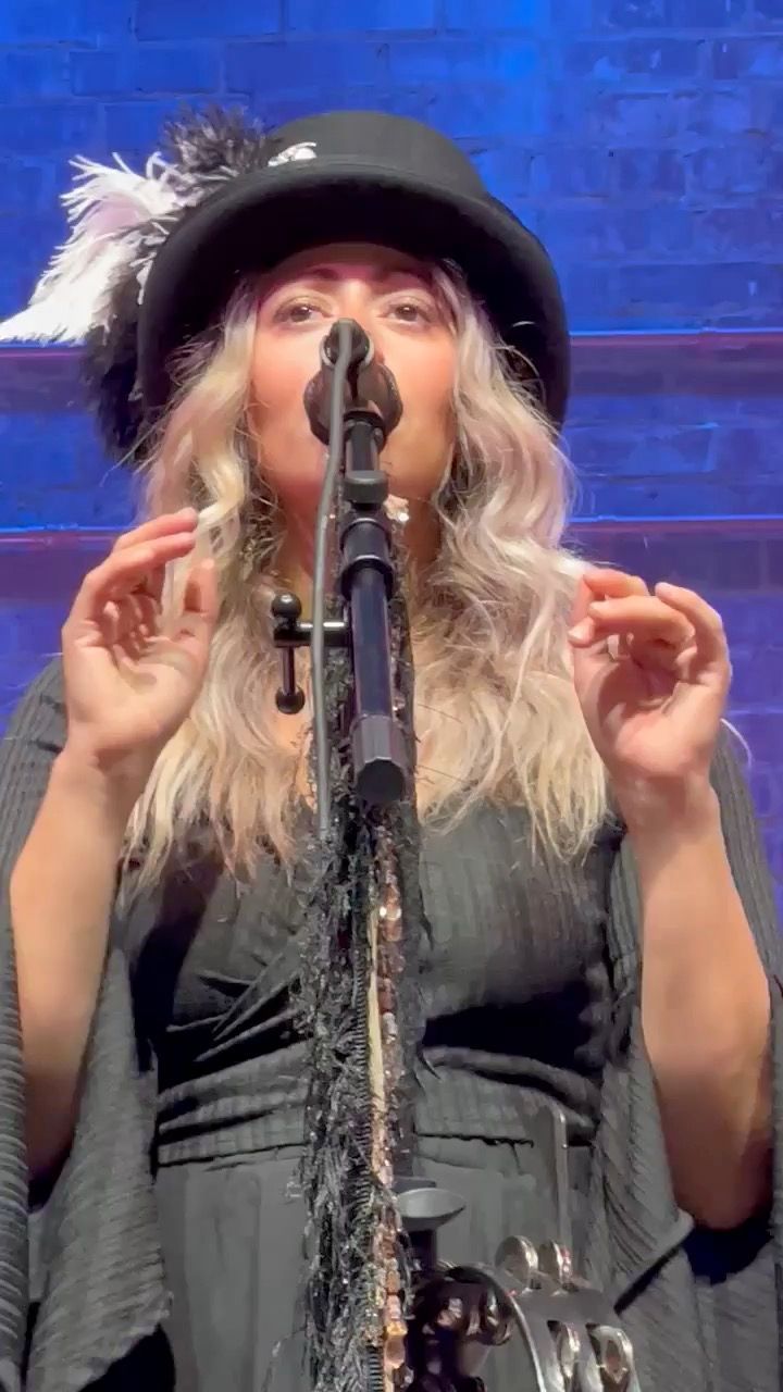 Woman with blonde hair wearing a hat with a feather, playing a saxophone and singing on stage.