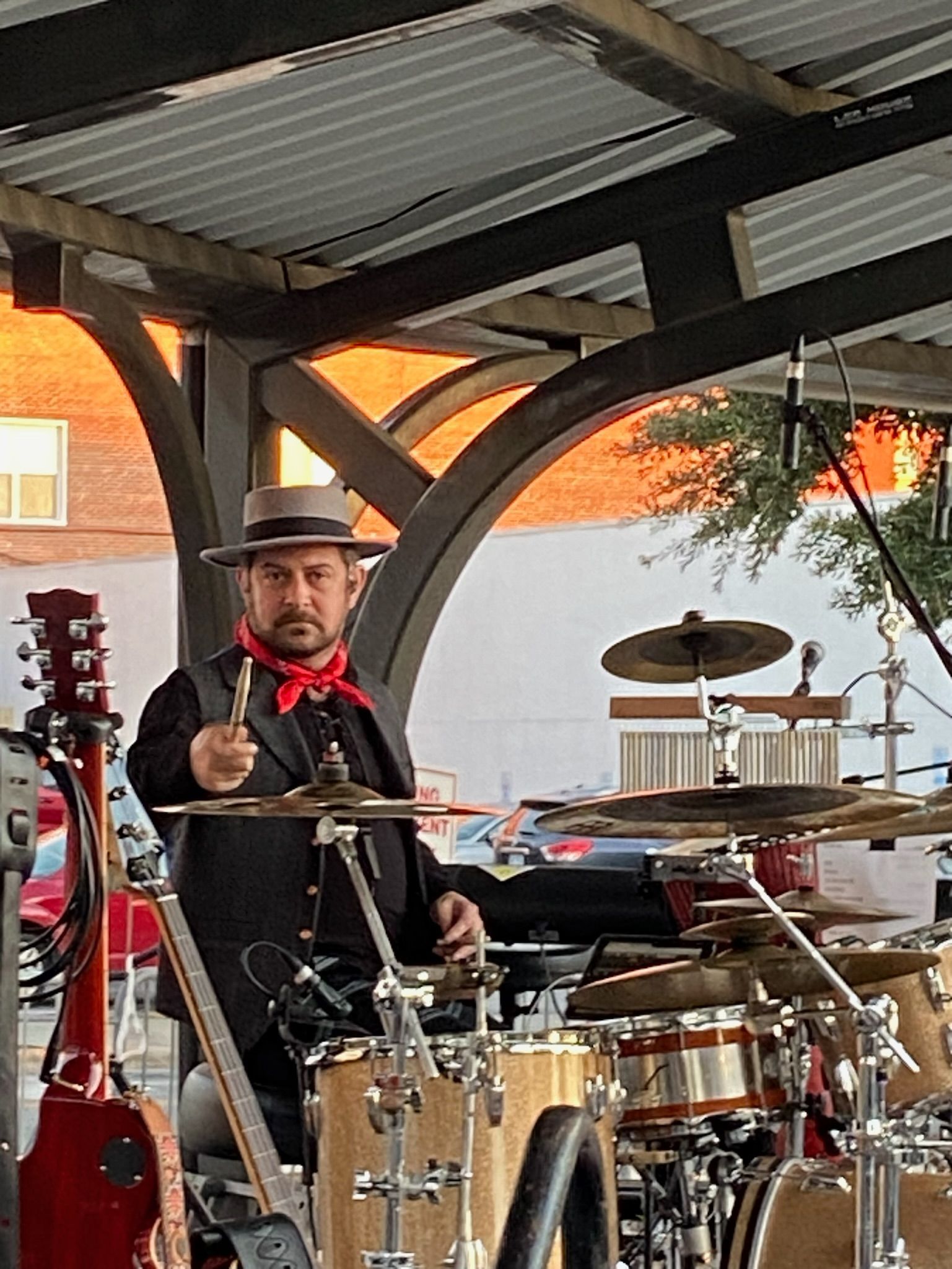 Drummer in hat and red scarf playing under an archway, drums visible.