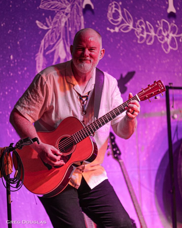 Man with a bald head and beard, playing an acoustic guitar onstage under purple lights.