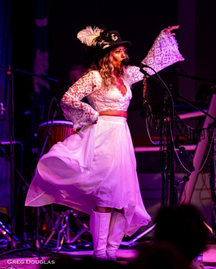 Woman in white lace, feathered hat, singing onstage, arm raised, purple and blue stage lighting.