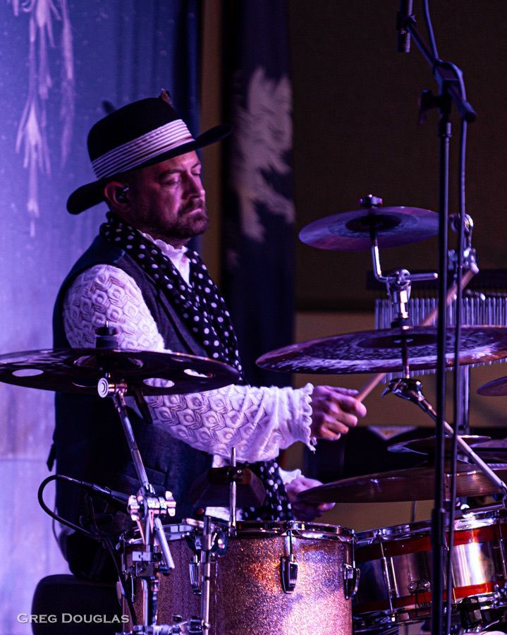 Drummer in a hat and vest playing at a concert. He is focused, illuminated by stage lights.