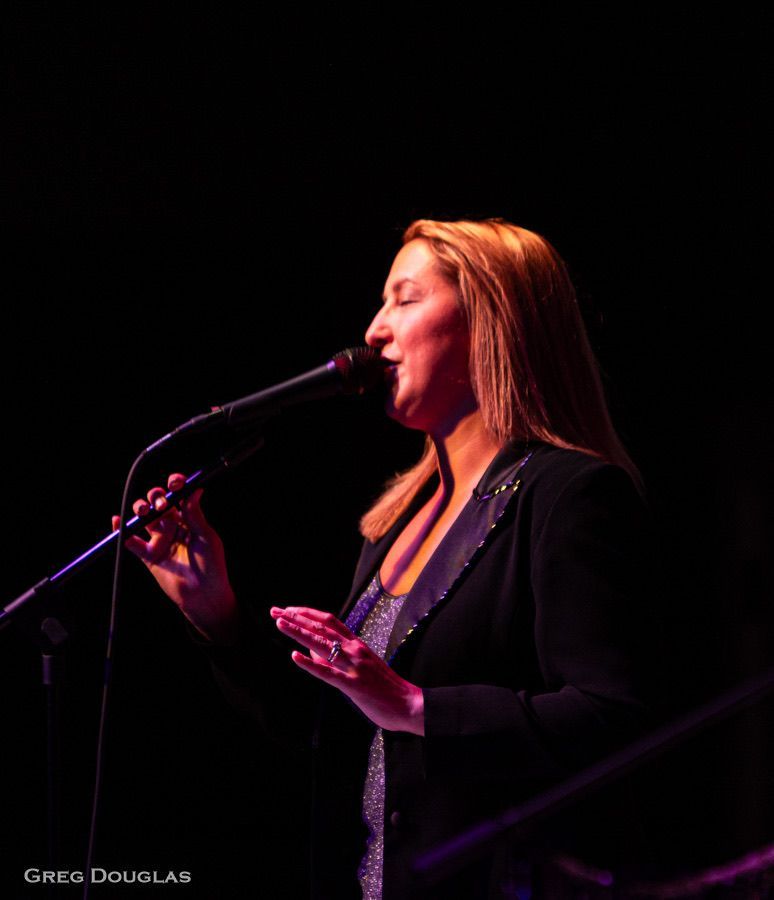 Woman singing into a microphone on stage, lit with stage lights, wearing a black blazer.