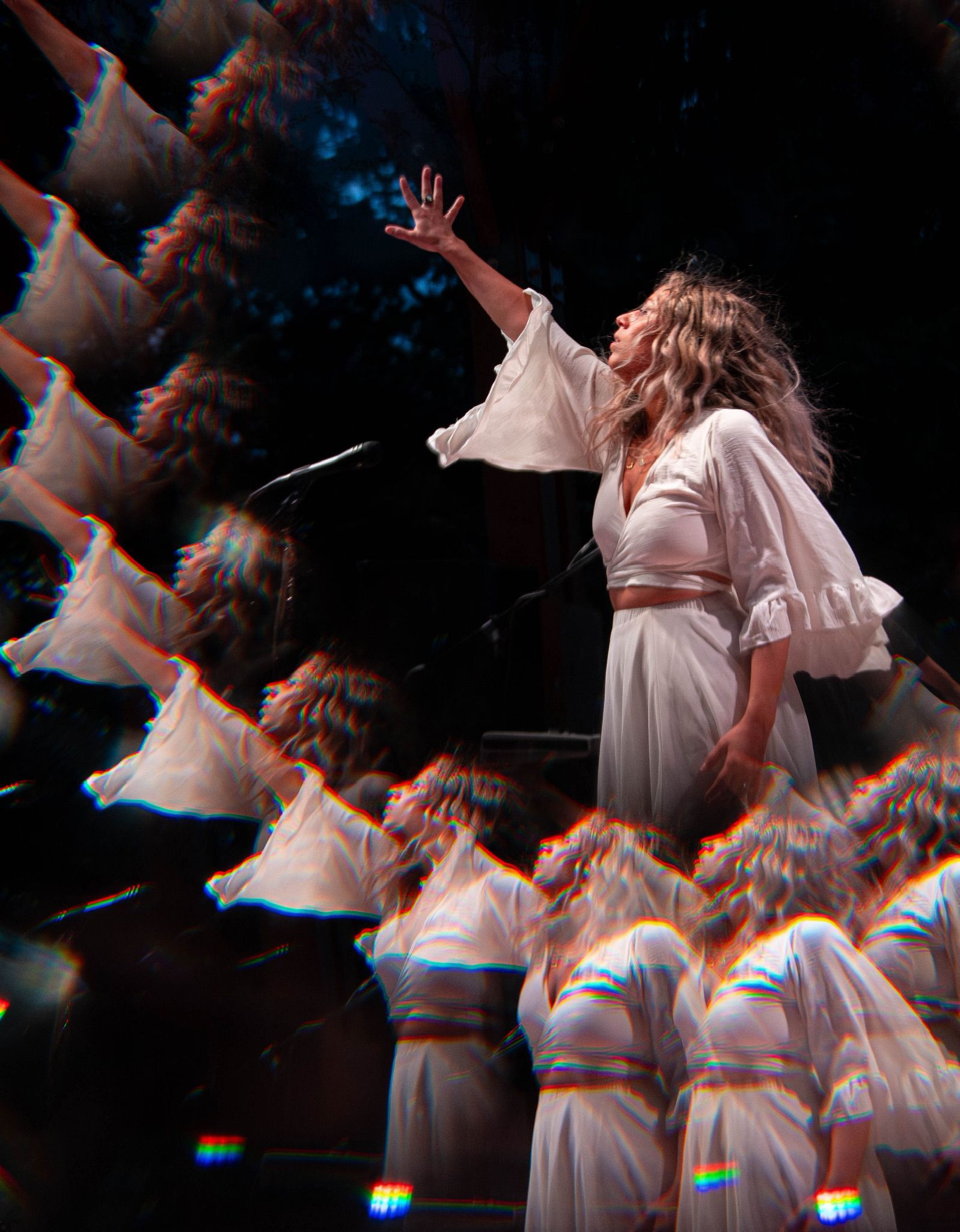 Woman in white dress reaching up, multiple reflections, dark background.