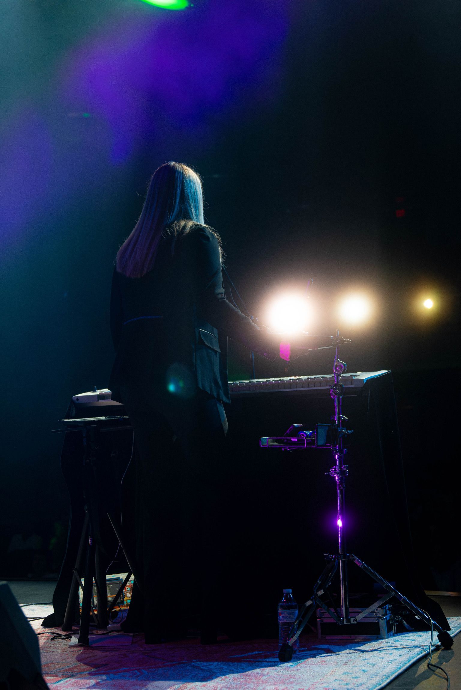 Person with long hair performing on stage, illuminated by spotlight at keyboard. Dark setting.