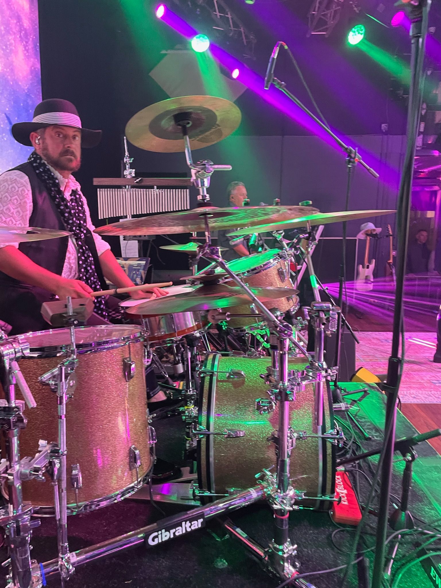 Drummer in a stage performance, playing a kit with gold drums and cymbals. Stage lights are green and purple.