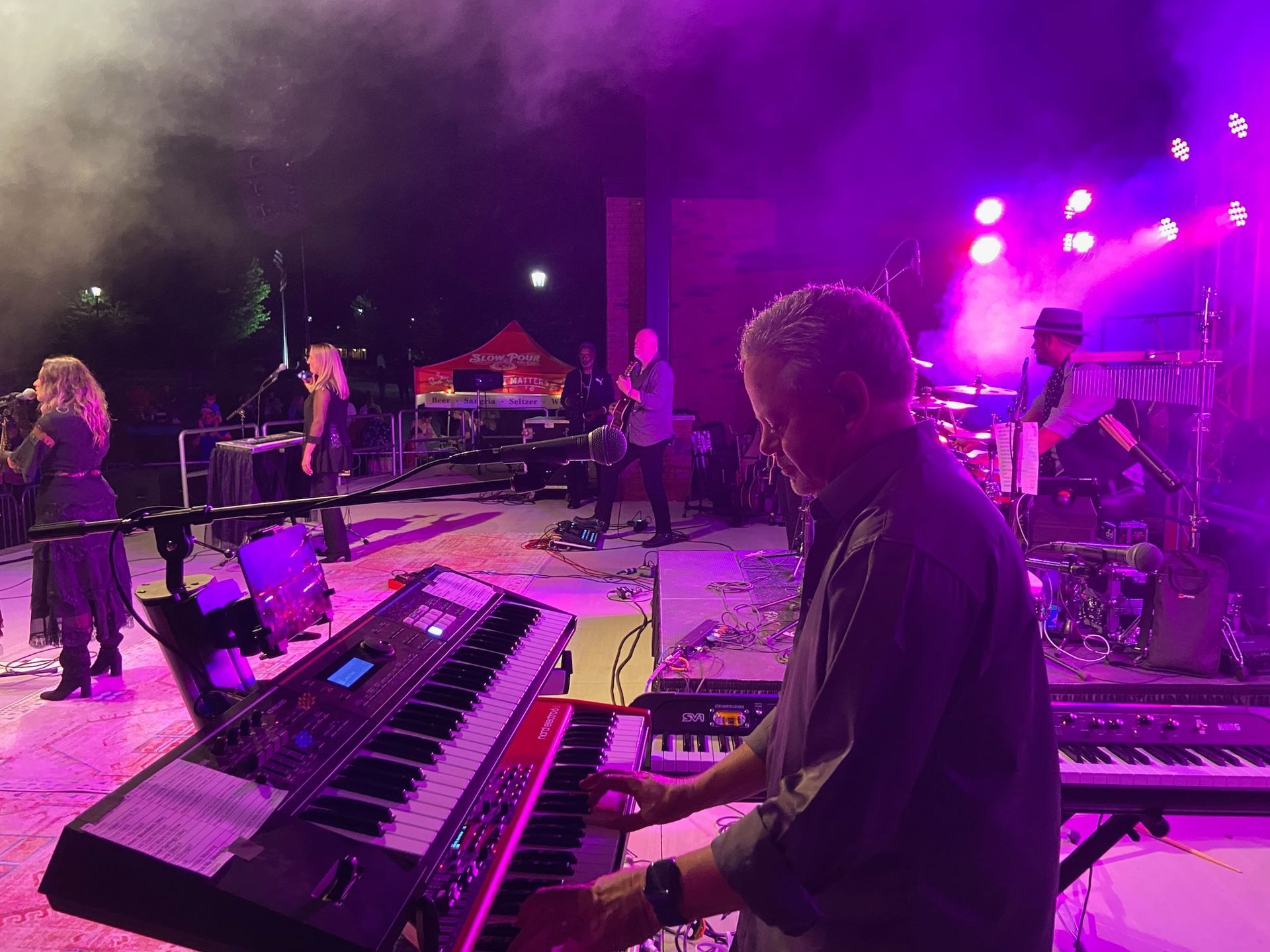 A band performing on an outdoor stage at night under purple lighting. Keyboardist in foreground, vocalists and other musicians behind.