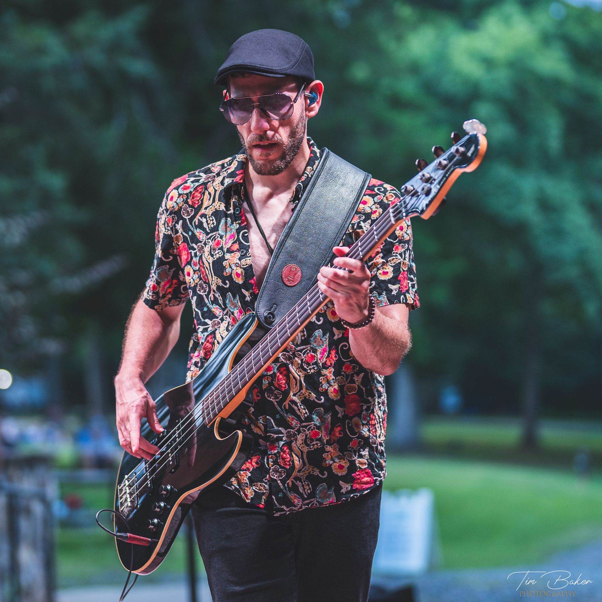 Man playing a black bass guitar on stage, wearing a patterned shirt, sunglasses, and a cap.