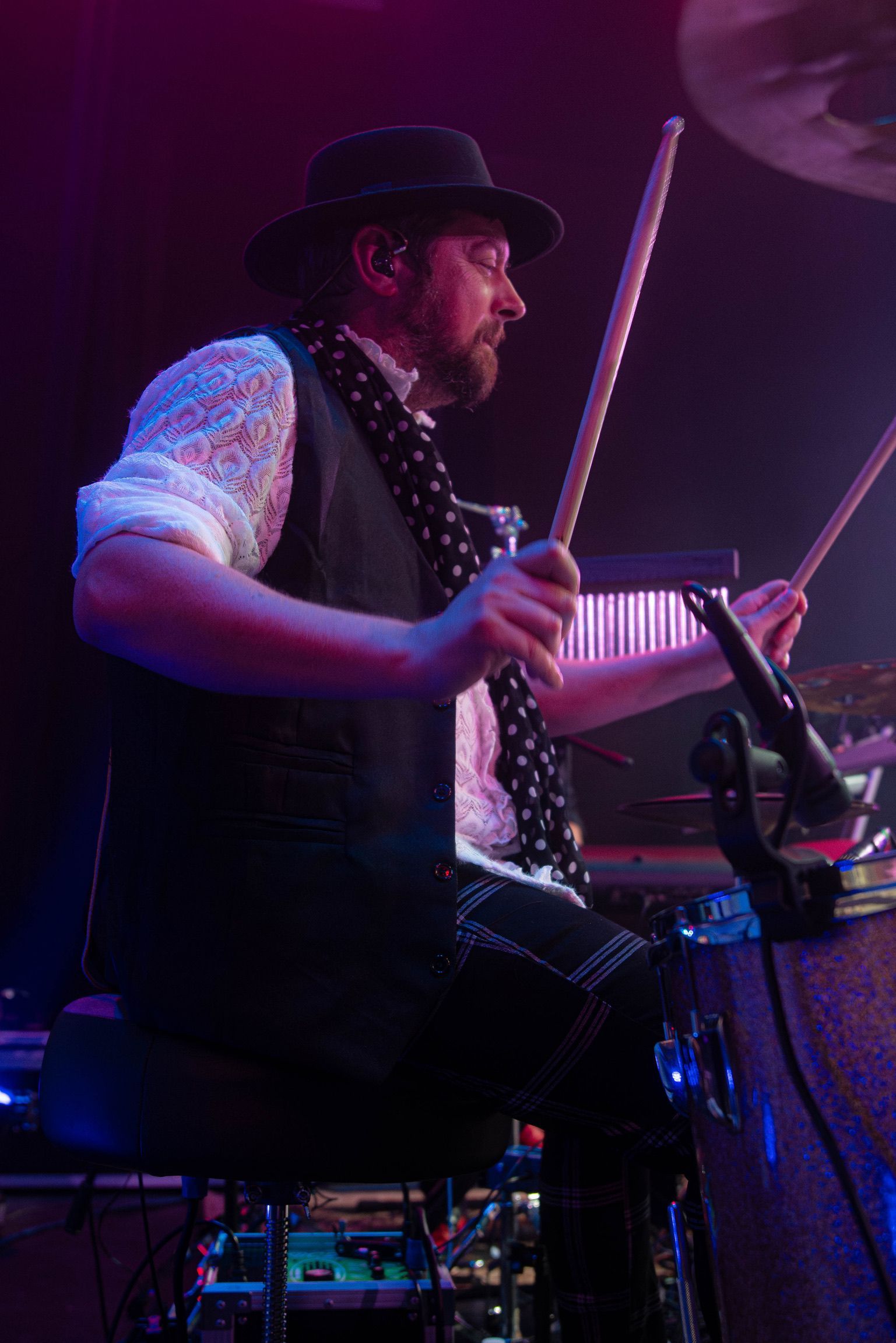 Drummer in a black hat, vest, and patterned scarf, playing drums on stage.