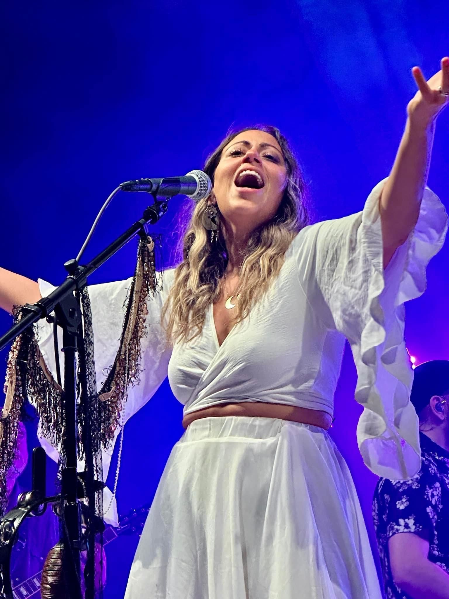 Singer on stage, arms raised, wearing a white dress, singing into a microphone, blue lighting.