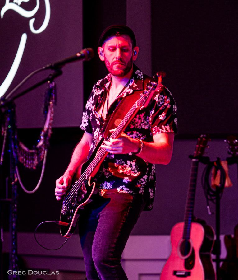 Man playing guitar on stage, wearing a floral shirt, dark hair, and jeans. Purple lighting.