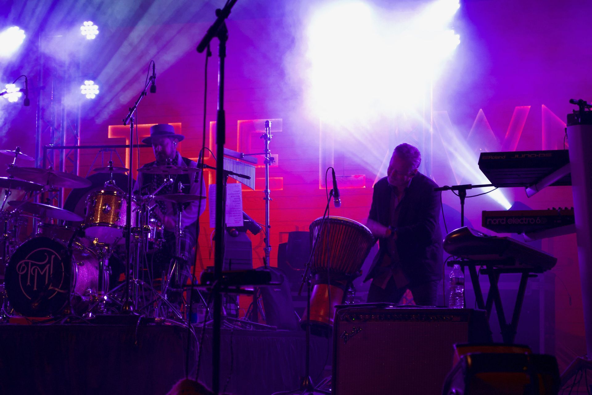A band performs on stage, bathed in purple and white stage lights. Drummer and percussionist are visible.