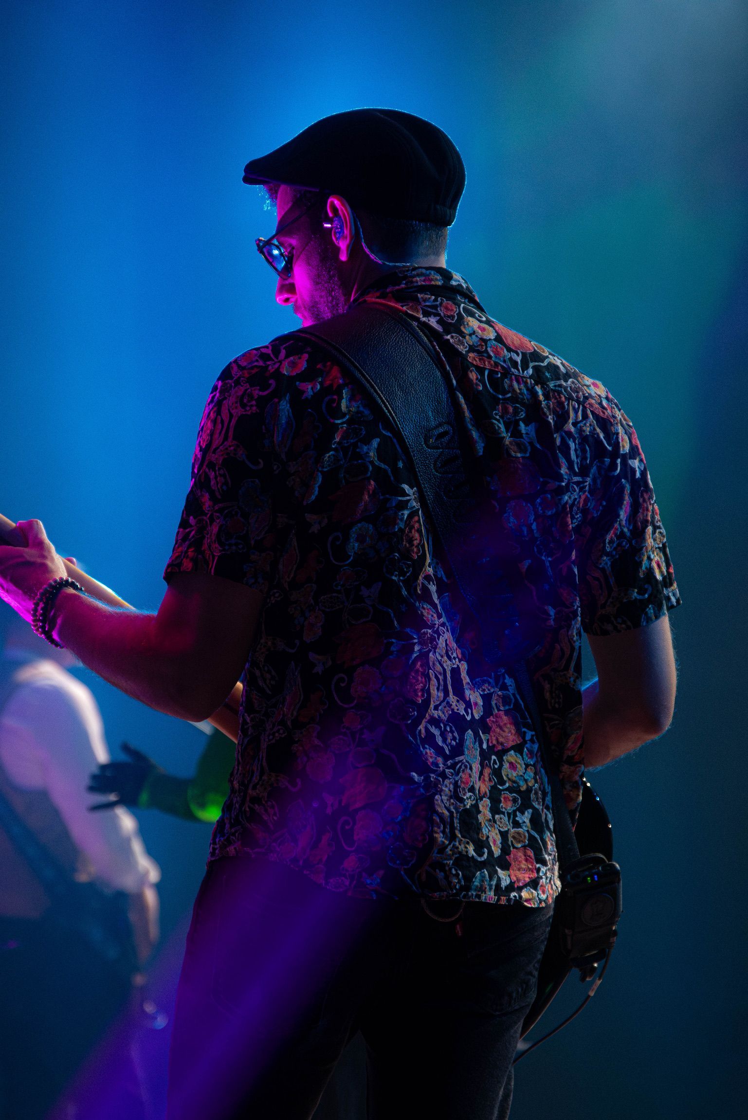 Man with guitar, wearing a patterned shirt and cap, onstage under blue and purple stage lights.