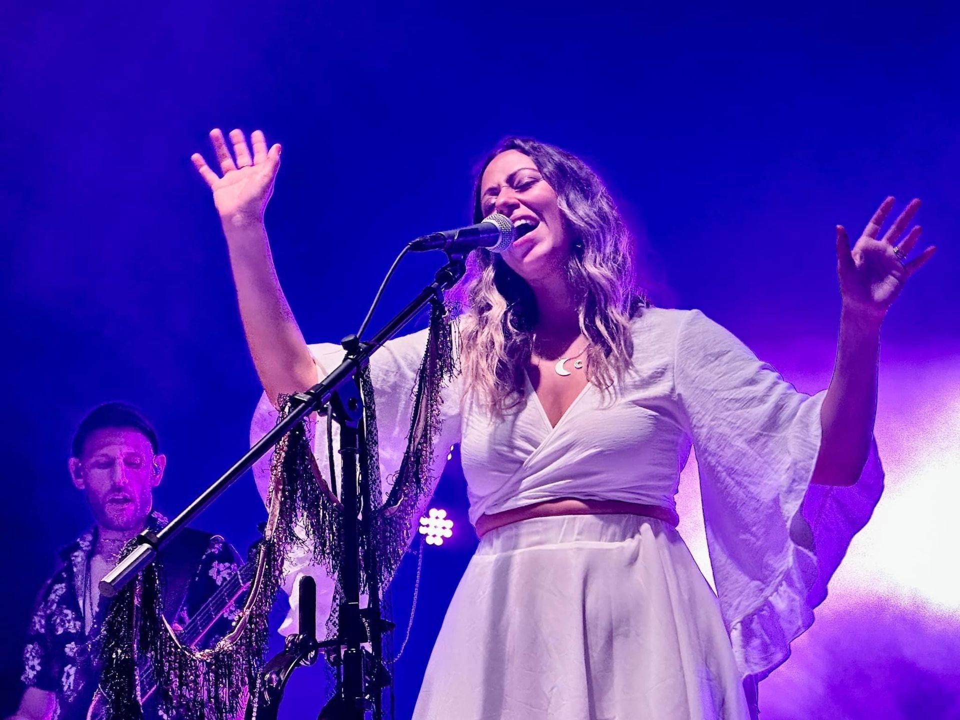 A woman in white singing on stage with arms raised; another musician is in the background.