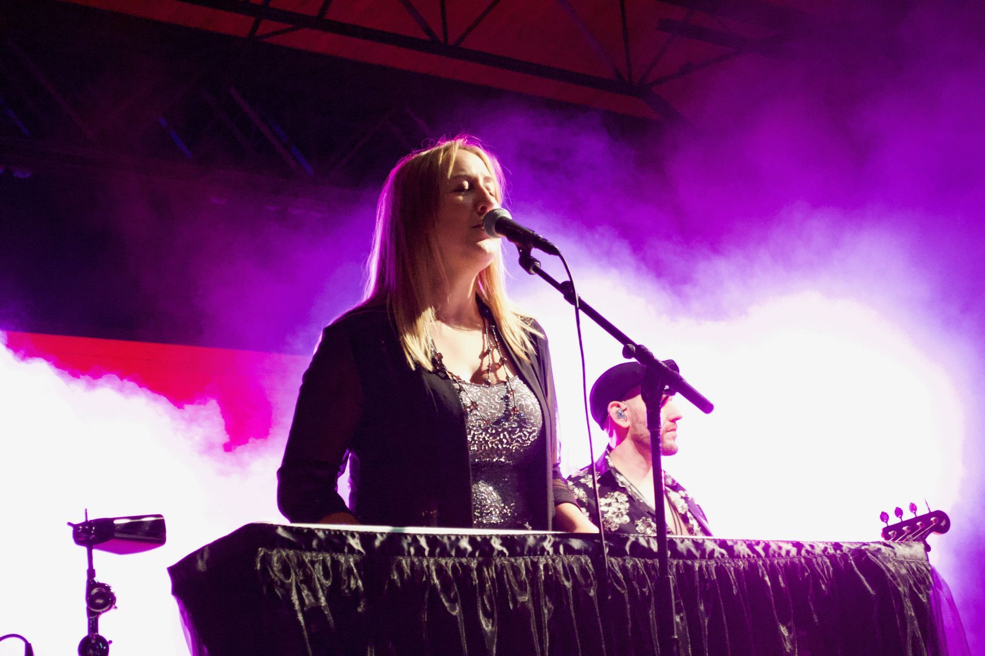 Woman singing at a concert, playing a keyboard with a man in background. Purple lighting and smoke.