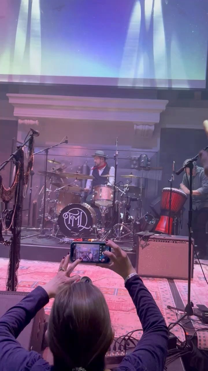Person taking photo of band on stage with a drummer, red rug, and stage lights.