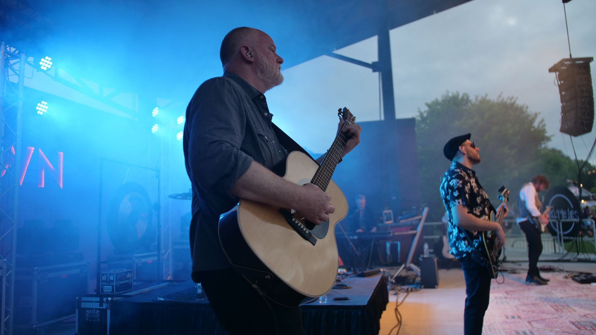 Band performing outdoors on a stage. Man with acoustic guitar in foreground, other band members behind. Blue stage lights.