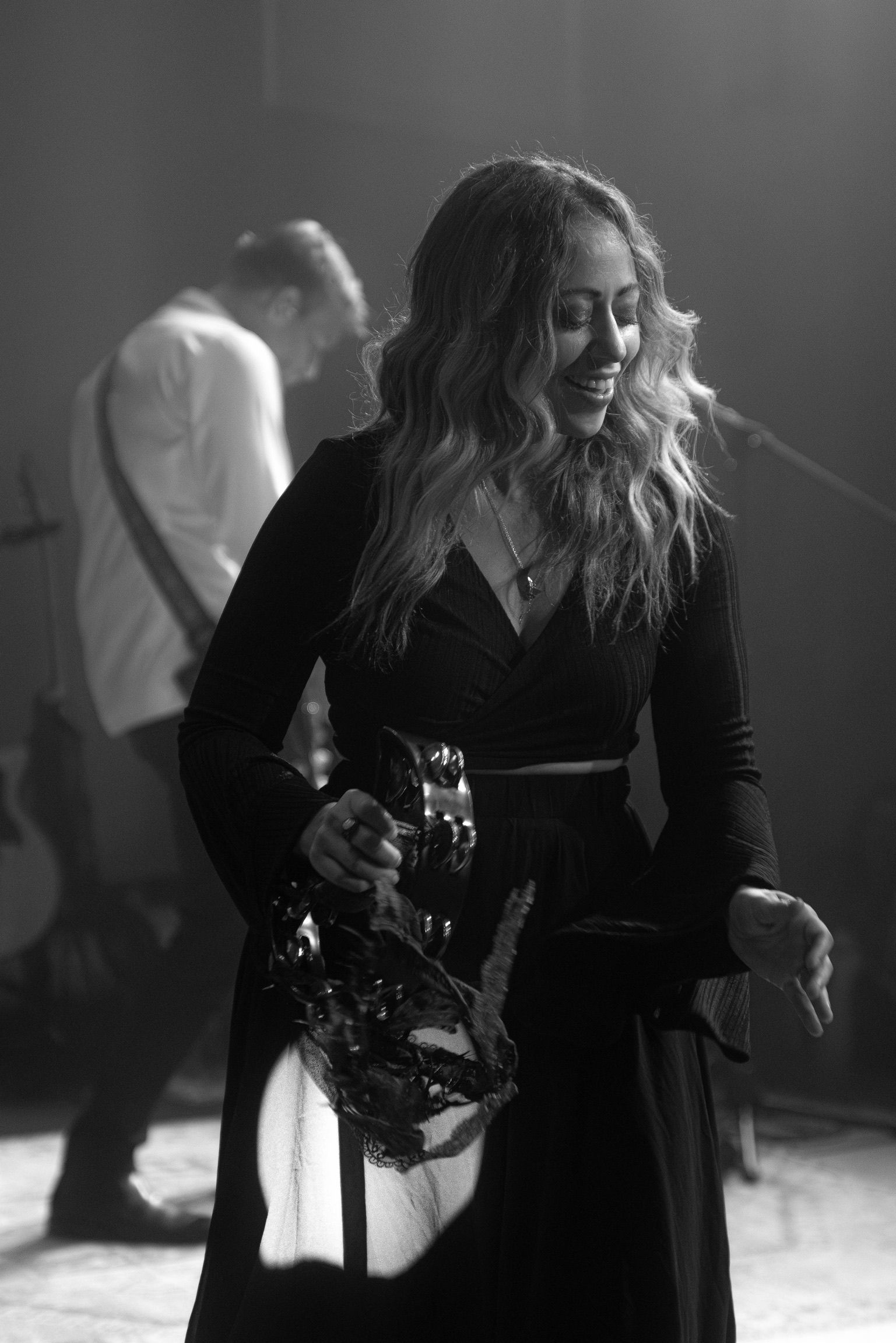 Woman smiling, holding instrument on stage, wearing black dress, with band member in the background.