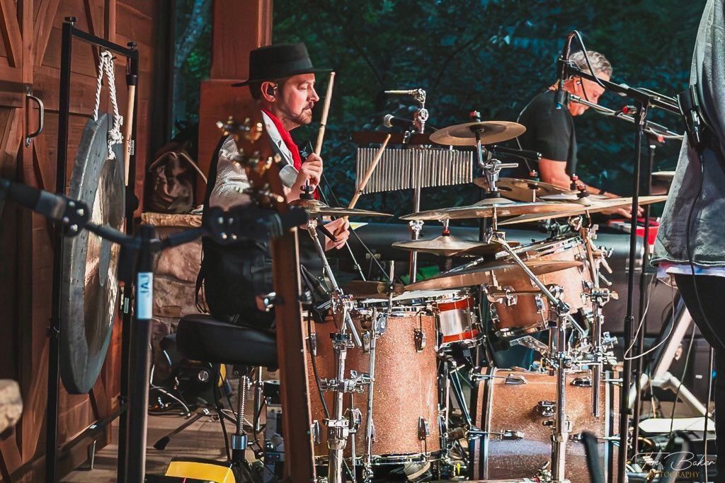 Drummer in a black hat performs on a drum set, with a gong and other percussion instruments, outdoor setting.