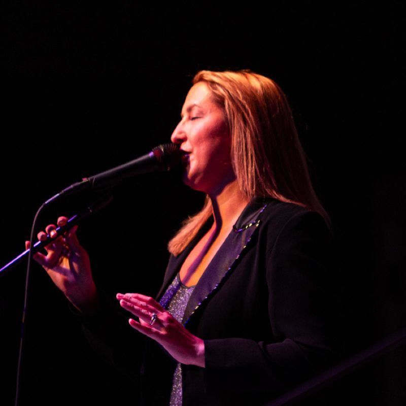Woman with blonde hair singing into a microphone on stage, lit by red and purple light.