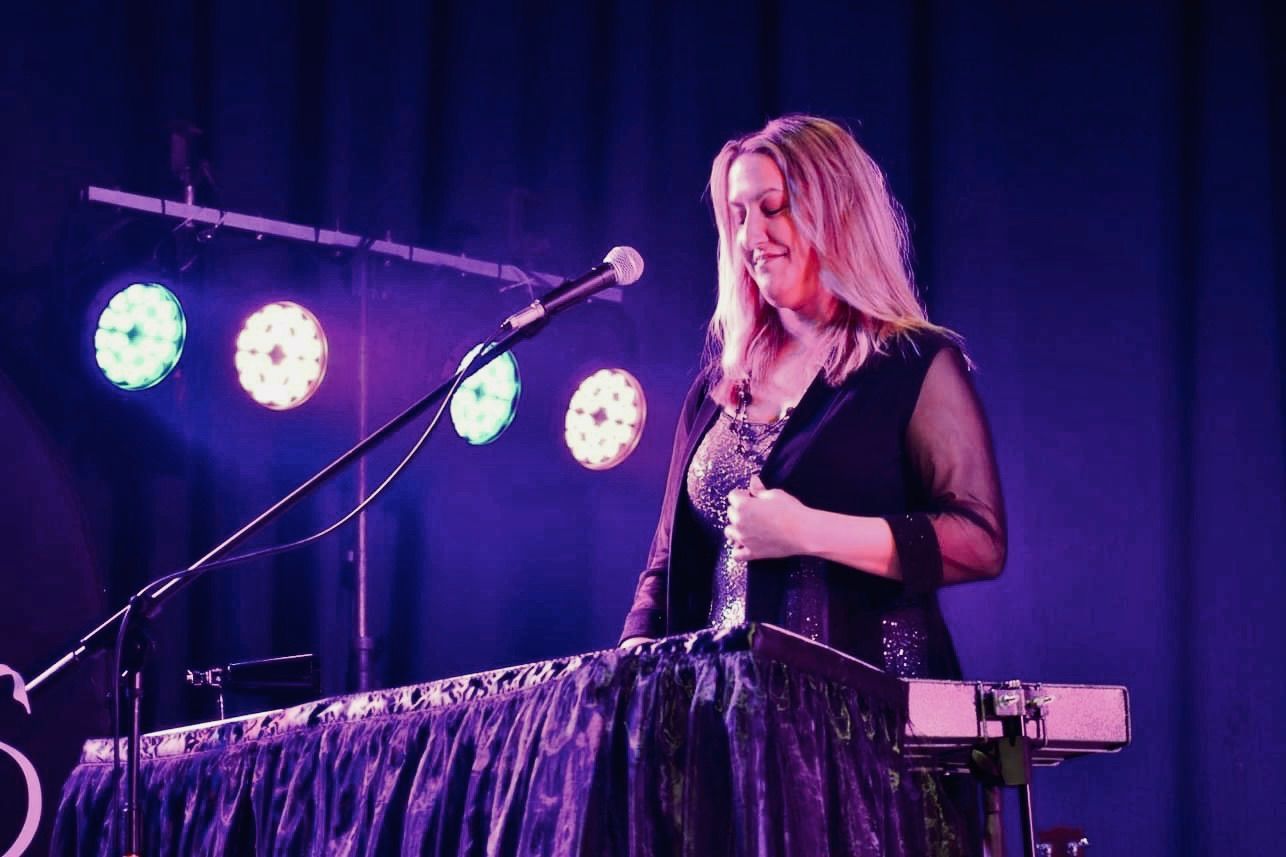 Woman playing keyboard on stage, lit by colorful spotlights.