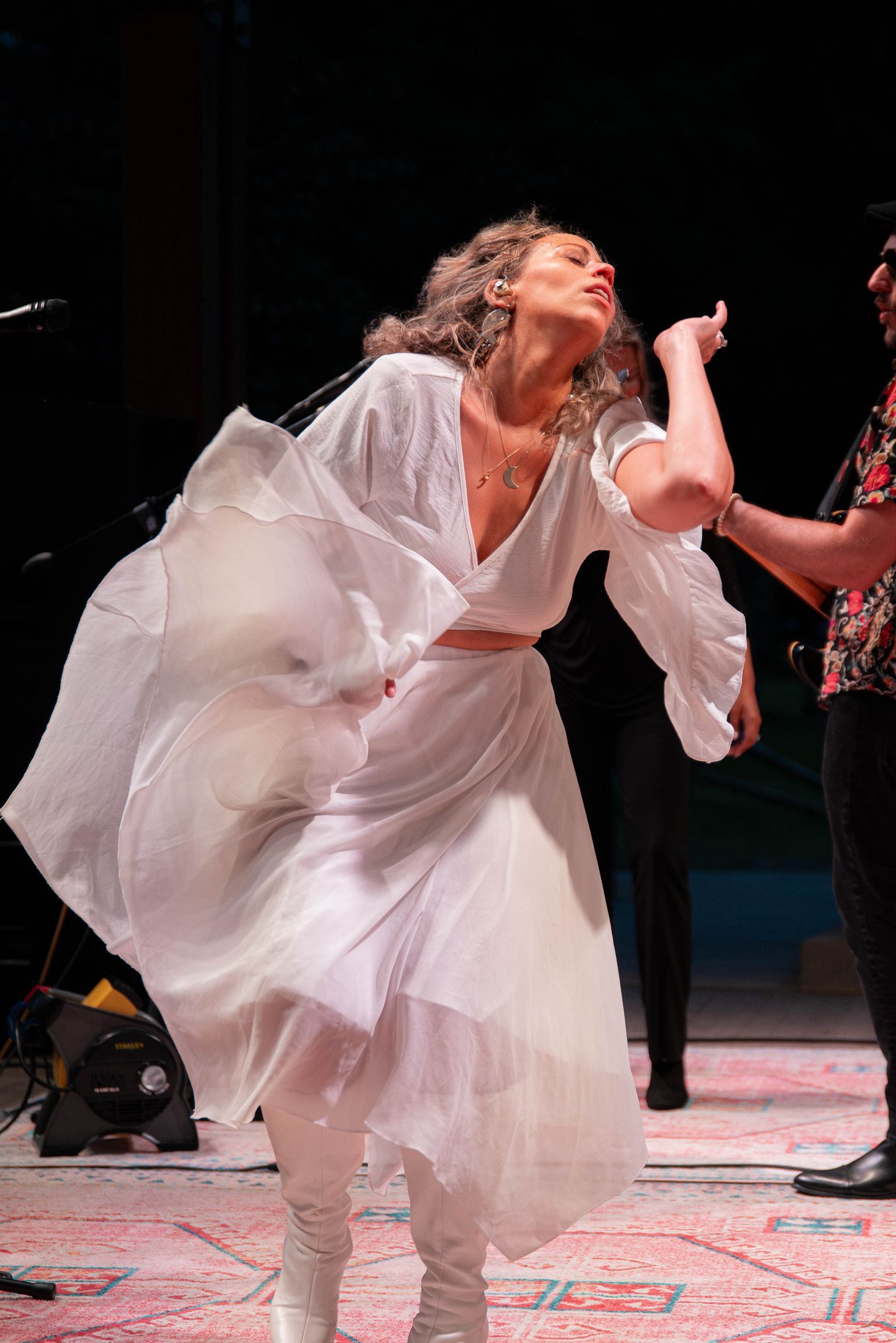 Woman in white dress dancing on stage, arms raised, light shining, with another person.