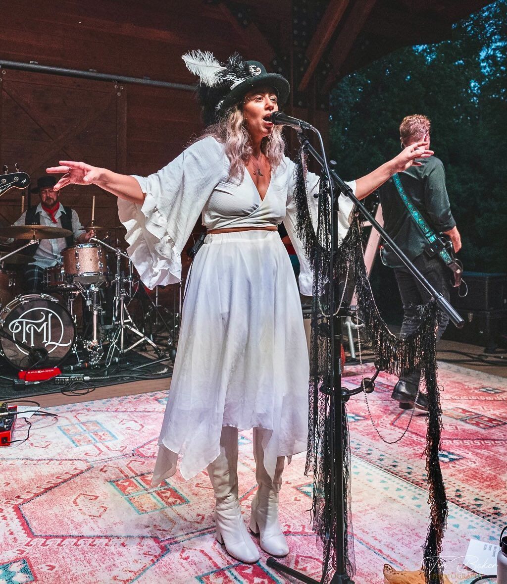 Woman singing onstage with a band, wearing white dress, hat with feathers, arms outstretched.