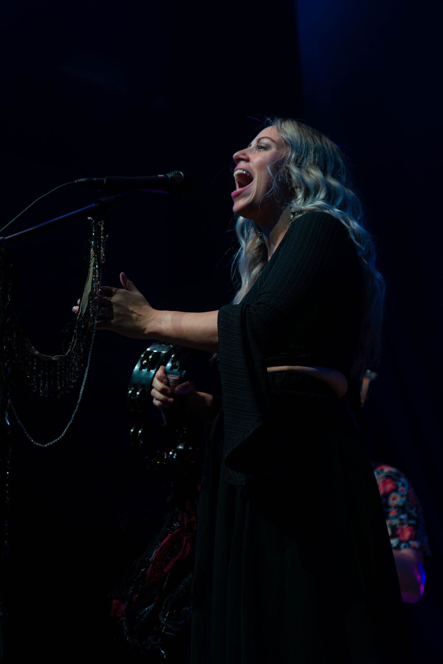 Woman on stage singing with saxophone, holding tambourine; dark setting.