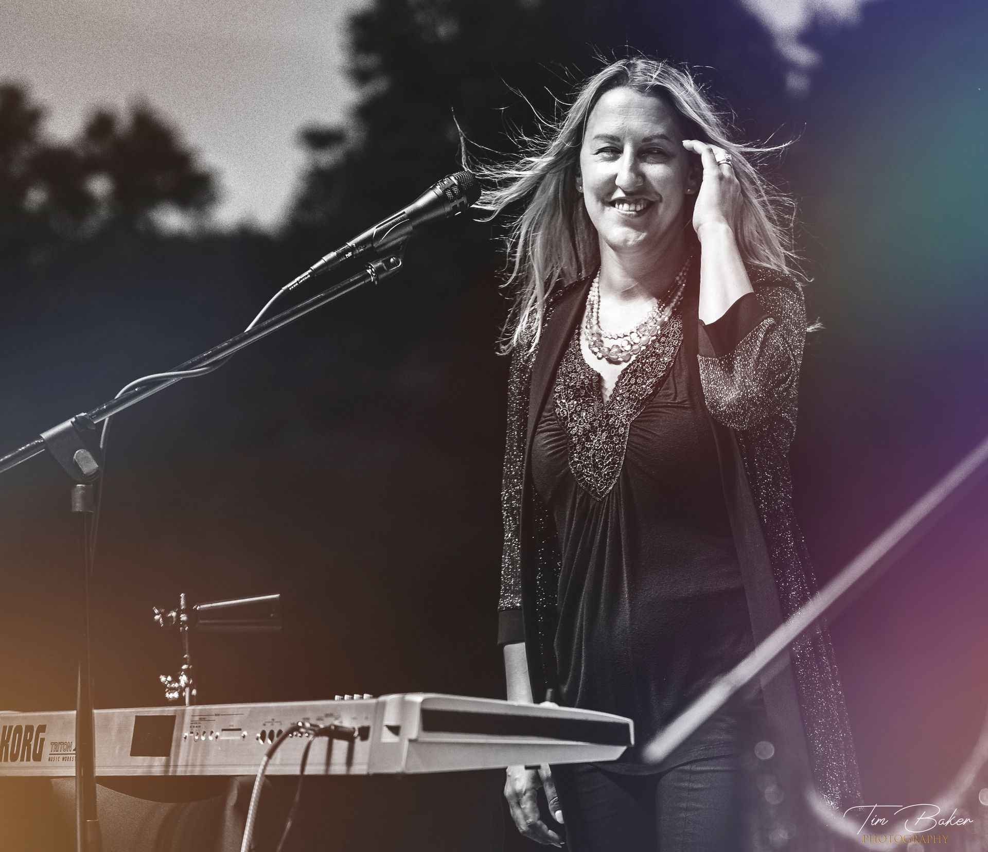 Woman playing keyboard onstage, smiling with hand in hair, wearing dark clothing with beaded jacket.