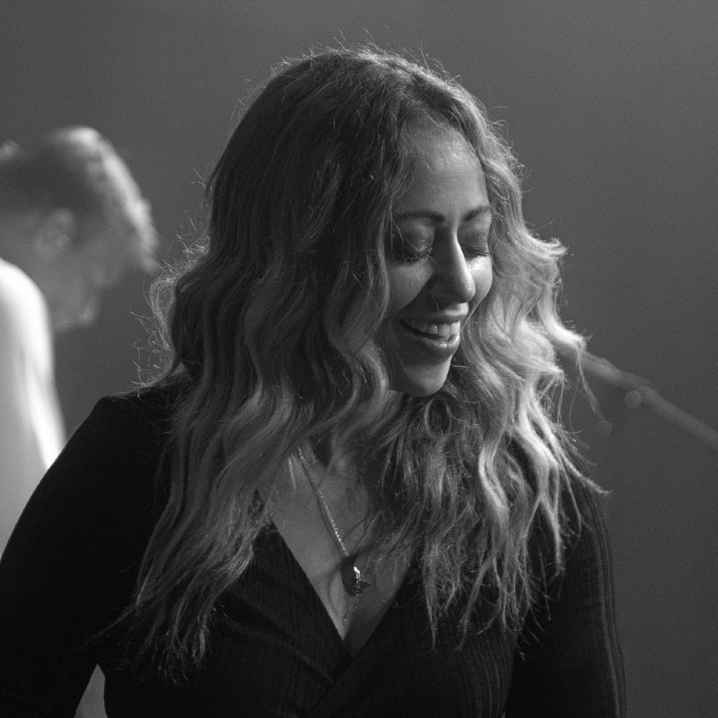 Woman with long wavy hair smiling and looking down, wearing a black top, on stage.