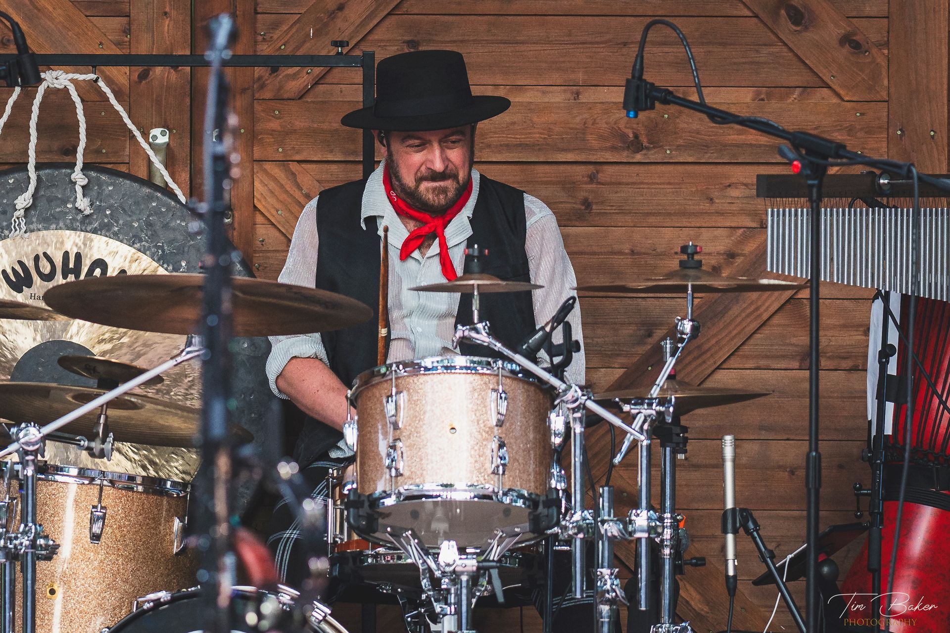 Drummer in a hat and vest, playing a kit in front of a wooden backdrop. Red scarf and cymbal.